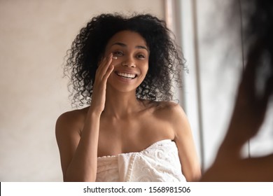 Smiling Young African American Woman In Towel Look In Mirror In Home Bathroom Apply Under Eye Cream, Happy Biracial Female Perform Daily Morning Facial Skincare Treatment, Use Serum, Beauty Concept