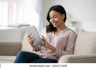 Smiling young African American woman sit relax on couch in living room browsing Internet shopping online on tablet, happy biracial female rest on sofa at home texting messaging on modern pad gadget