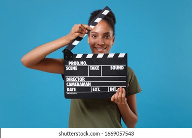 Smiling young african american woman girl in casual t-shirt posing isolated on bright blue background. People lifestyle concept. Mock up copy space. Holding classic black film making clapperboard