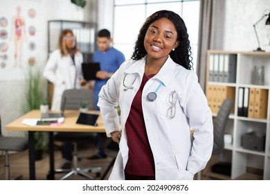 Smiling Young African American Woman Nurse Standing In Bright Modern Hospital Meeting Room In Front Of Two Co-workers, Discussing On The Background