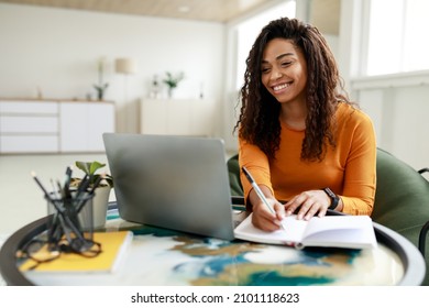 Smiling young African American woman sitting at desk working on laptop taking notes in notebook, happy millennial female studying online, watching webinar using computer and writing check list