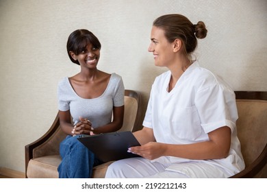 Smiling Young African American Woman Visiting Doctor In Clinic. Happy Female Doctor Or Nurse Wearing Lab Coat, Holding Clipboard And Talking To Female Patient. Visiting Doctor Concept