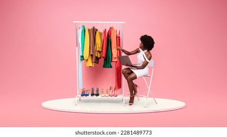 Smiling young african american woman fashion designer in white dress with laptop looks at clothes and shoes on platform in showroom on pink studio background. Fashion, style and modern device - Powered by Shutterstock