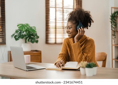 Smiling young african american woman in wireless headphones sitting at desk table working on laptop and writing letter in paper notebook, taking notes watching weninar - Powered by Shutterstock