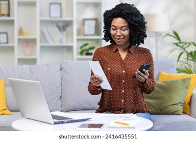 Smiling young African American woman sitting at home on couch at table with laptop, paying and paying bills via phone, holding documents, receipts. - Powered by Shutterstock