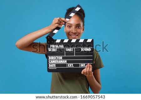 Smiling young african american woman girl in casual t-shirt posing isolated on bright blue background. People lifestyle concept. Mock up copy space. Holding classic black film making clapperboard
