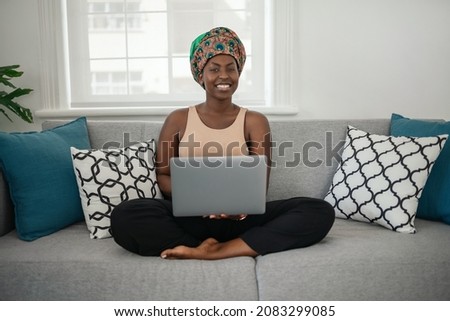 Smiling young African American woman sitting with her legs crossed on her sofa at home using laptop and looking into camera