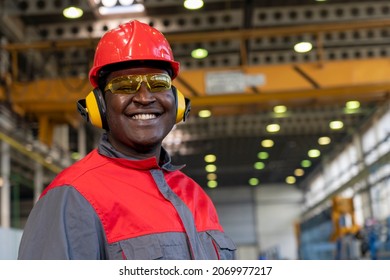 Smiling Young African American Worker In Personal Protective Equipment Looking At Camera. Portrait Of Black Industrial Worker In Red Helmet, Hearing Protection Equipment And Work Uniform In A Factory. - Powered by Shutterstock