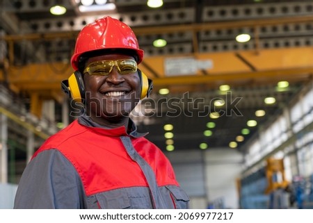 Smiling Young African American Worker In Personal Protective Equipment Looking At Camera. Portrait Of Black Industrial Worker In Red Helmet, Hearing Protection Equipment And Work Uniform In A Factory.