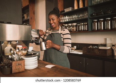 Smiling young African barista frothing milk for a cappuccino behind the counter of her trendy cafe - Powered by Shutterstock