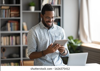Smiling young african business man professional holding using digital tablet computer online app software on tech device modern pad gadget browse internet work communicate online stand at home office