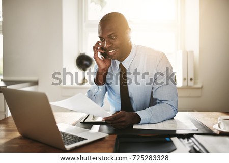 Smiling young African businessman talking to a client on his cellphone while sitting at his desk in an office