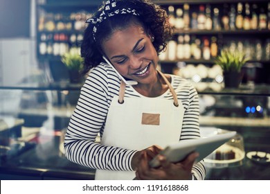 Smiling Young African Cafe Owner Taking Reservations Over The Phone And Using A Digital Tablet