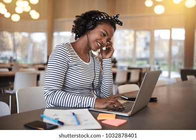 Smiling young African college student sitting on campus working online with a laptop and listening to music on headphones - Shutterstock ID 1032878401