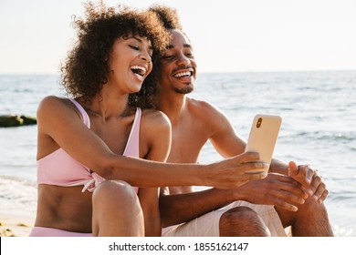 Smiling young african couple sitting together on a sandy beach taking selfies