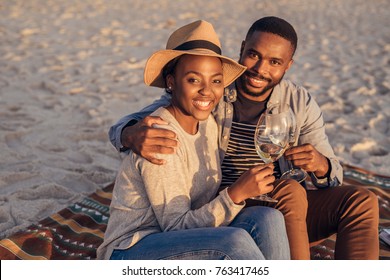 Smiling young African couple sitting on a sandy beach enjoying a glass of wine while watching a sunset - Powered by Shutterstock