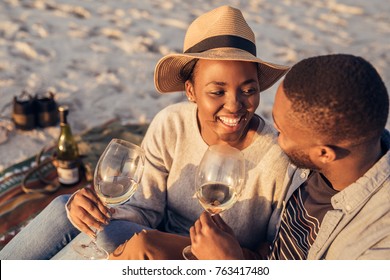 Smiling Young African Couple Sitting Together On A Sandy Beach Enjoying A Glass Of Wine And A Late Afternoon Picnic