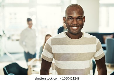 Smiling young African designer standing in a modern office with a group of colleagues working in the background