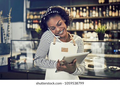 Smiling young African entrepreneur standing in front of the counter of her cafe talking on a cellphone and using a tablet