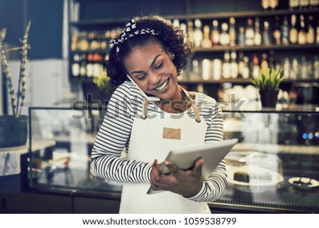 Smiling young African entrepreneur standing in front of the counter of her cafe talking on a cellphone and using a tablet