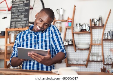 Smiling young African entrepreneur talking on a cellphone and using a digital tablet while standing at the counter of his cafe
