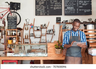 Smiling young African entrepreneur wearing an apron leaning against the counter of his trendy cafe using a digital tablet