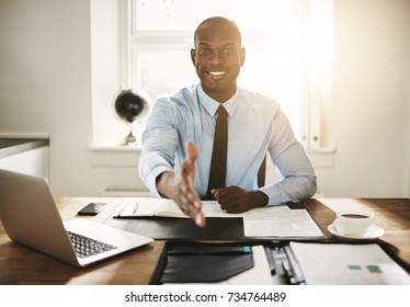 Smiling young African executive extending his arm in a handshake while sitting at his desk in an office