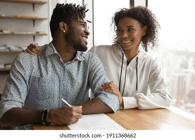 Smiling young african family couple clients customers of real estate agency embrace talk when buy property take up mortgage loan. Happy black spouses support one another in signing contract with bank