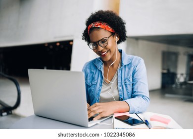 Smiling young African female entrepreneur using a laptop while working at a table in the lobby of a modern office building