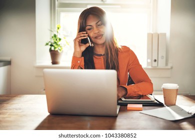 Smiling Young African Female Entrepreneur Talking On A Cellphone And Working On A Laptop While Sitting At A Table In Her Home Office