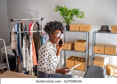 Smiling young African female entrepreneur working on a tablet PC while sitting at a table in her home office. Young woman talking on a cellphone while working on her tablet PC at home - Powered by Shutterstock