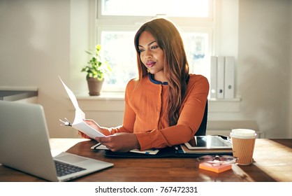 Smiling Young African Female Entrepreneur Sitting At A Table In Her Home Office Reading Paperwork And Working On A Laptop