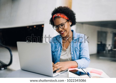 Smiling young African female entrepreneur using a laptop while working at a table in the lobby of a modern office building