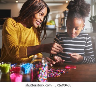 Smiling Young African Female Parent Helping Cute Child In Striped Shirt Make A Beaded Craft Heart Shape On Table In Kitchen