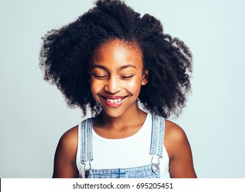 Smiling young African girl with long curly hair and wearing dungarees standing with her eyes closed against a gray background