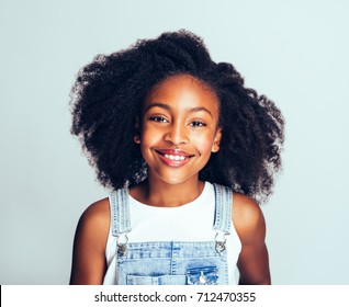 Smiling young African girl with long curly hair wearing dungarees standing happily against a gray background