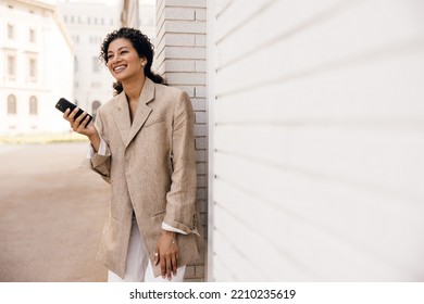 Smiling Young African Girl Spends Time On Europe City Street With Her Smartphone In Sunny Weather. Model With Brunette Hair Wears Business Style Of Clothing. Concept Modern Lifestyle.