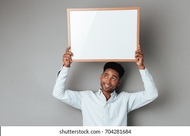 Smiling Young African Man Holding Blank Board Above His Head Isolated Over Gray Background