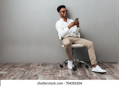 Smiling Young African Man Sitting In A Chair And Holding Mobile Phone Isolated Over Gray Background