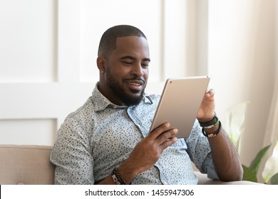 Smiling Young African Man Using Digital Tablet At Home, Millennial Black Male User Holding Computer Looking At Screen Reading E-book App Online Relaxing On Leisure With Device Work Study Sit On Sofa