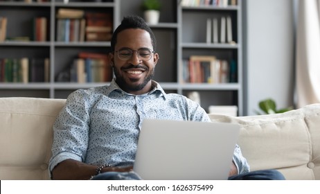 Smiling Young African Man Using Laptop Notebook Surfing Social Media Watching Movies Relax At Home On Couch, Happy Ethnic Guy Enjoy Easy E Learning Browse Internet On Modern Tech Device Sit On Sofa