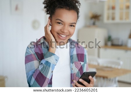 Smiling young African teen girl listens to music in wireless earphone holding smartphone, using mobile apps at home. Happy biracial teen lady enjoy sound, listening to audio book or podcast indoors