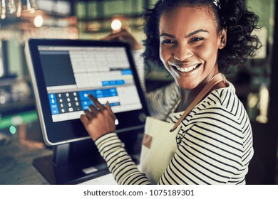 Smiling young African waitress wearing an apron working in a trendy restaurant and using a touchscreen point of sale terminal