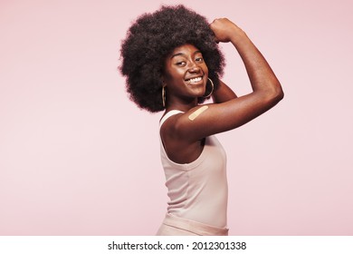 Smiling young African woman flexing her arm with a bandage on it after a Covid vaccination against a pink background - Shutterstock ID 2012301338