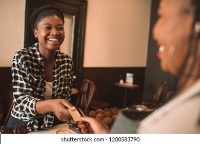 Smiling Young African Woman Giving Her Bank Card To A Cafe Cashier Pay For Her Cappuccino