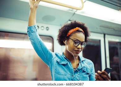 Smiling young African woman listening to music on her cellphone while standing on a subway train during her daily commute - Powered by Shutterstock