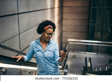 Smiling young African woman listening to music on her cellphone while riding alone up an escalator in a modern building - Powered by Shutterstock