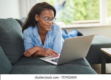 Smiling young African woman lying down on her living room sofa using a laptop to browse the internet