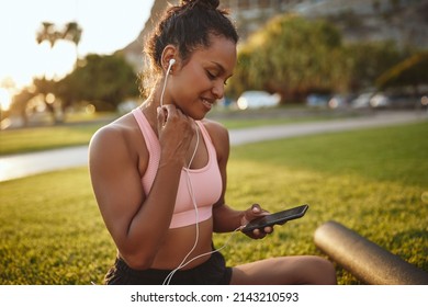 Smiling young African woman in sportswear sitting in a park listening to music on earphones after a yoga session - Shutterstock ID 2143210593