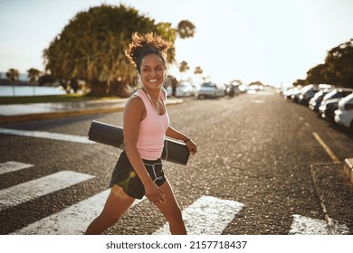 Smiling young African woman in sportswear crossing a street carrying a yoga mat and listening to music on earphones  - Powered by Shutterstock